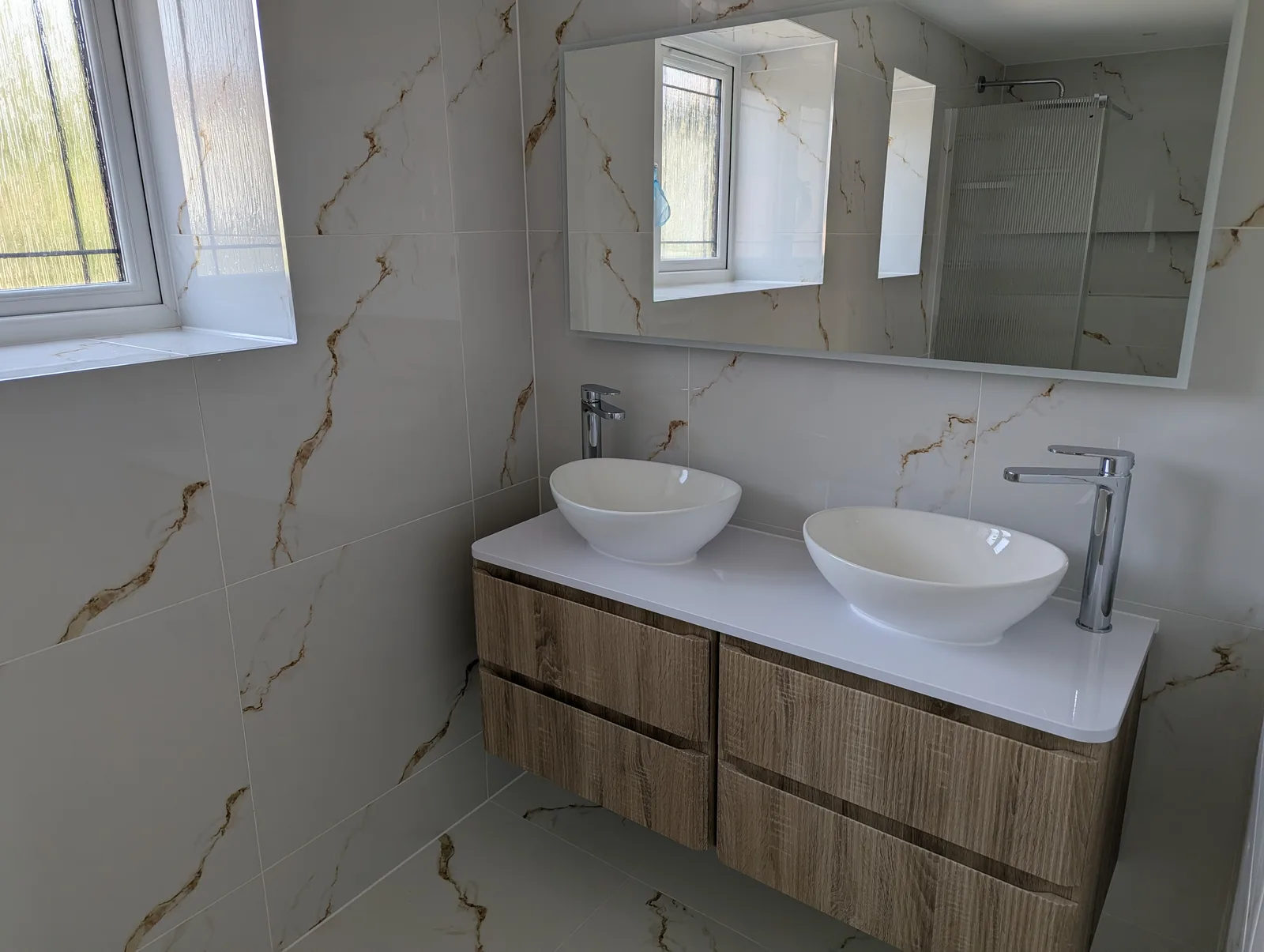 Twin countertop basins on a wall-hung oak-effect vanity unit with chrome tall taps and a full-width mirror, against calacatta-effect tiling