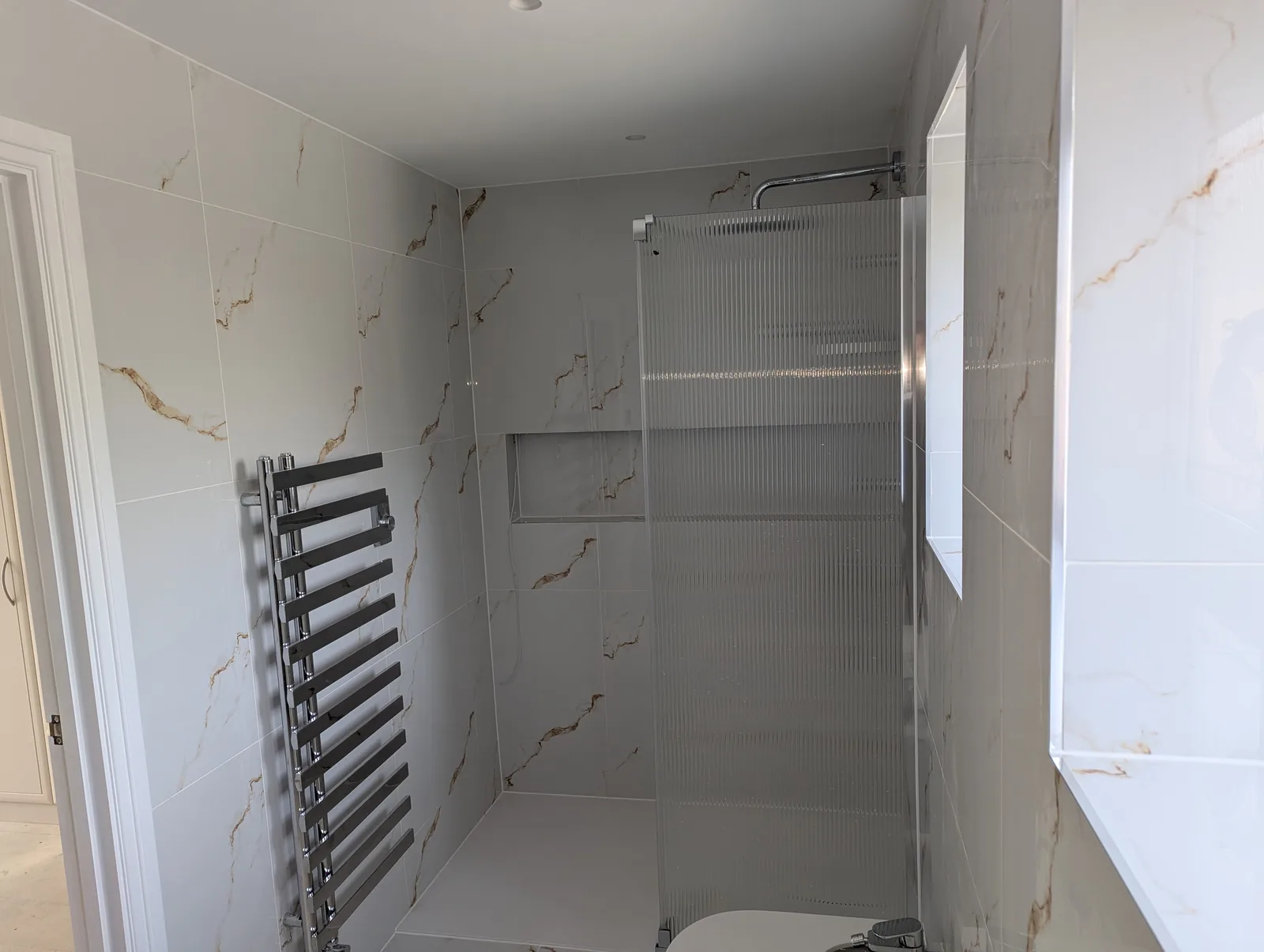 Walk-in shower with a low-profile tray, reeded glass screen, recessed tiled niche and chrome ladder radiator, in calacatta-effect porcelain