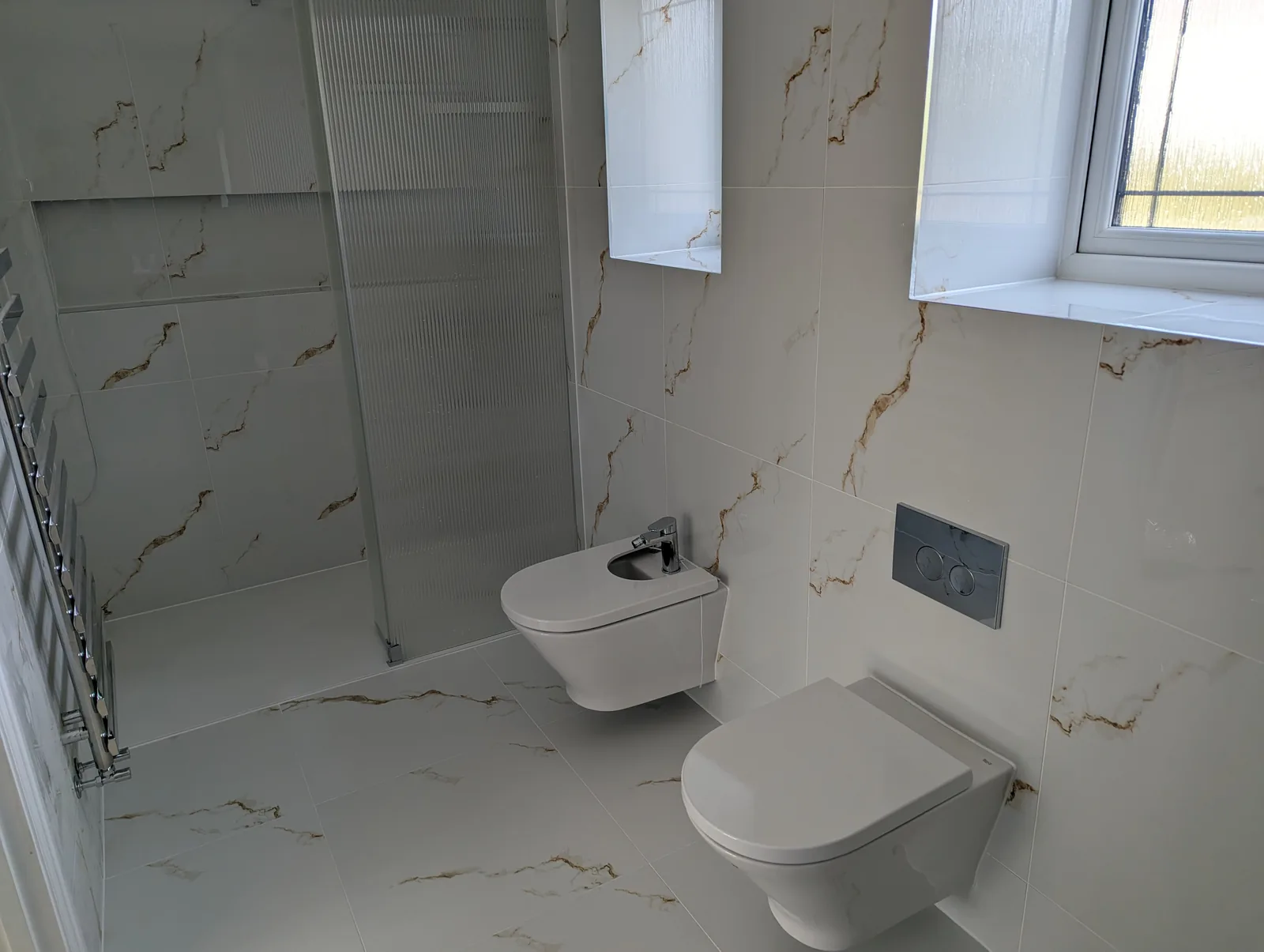 Wall-hung toilet and bidet with a concealed cistern and chrome flush plate, beside a reeded glass shower screen